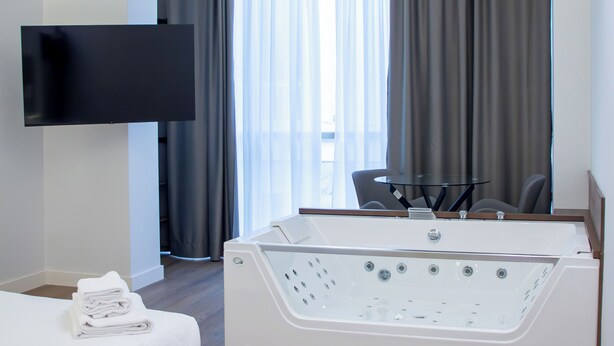 Une chambre d’hôtel moderne dotée d’une grande baignoire, d’un téléviseur à écran plat fixé au mur, d’un lit avec des serviettes soigneusement pliées et offrant une vue sur l’extérieur grâce à de grandes fenêtres équipées de rideaux.