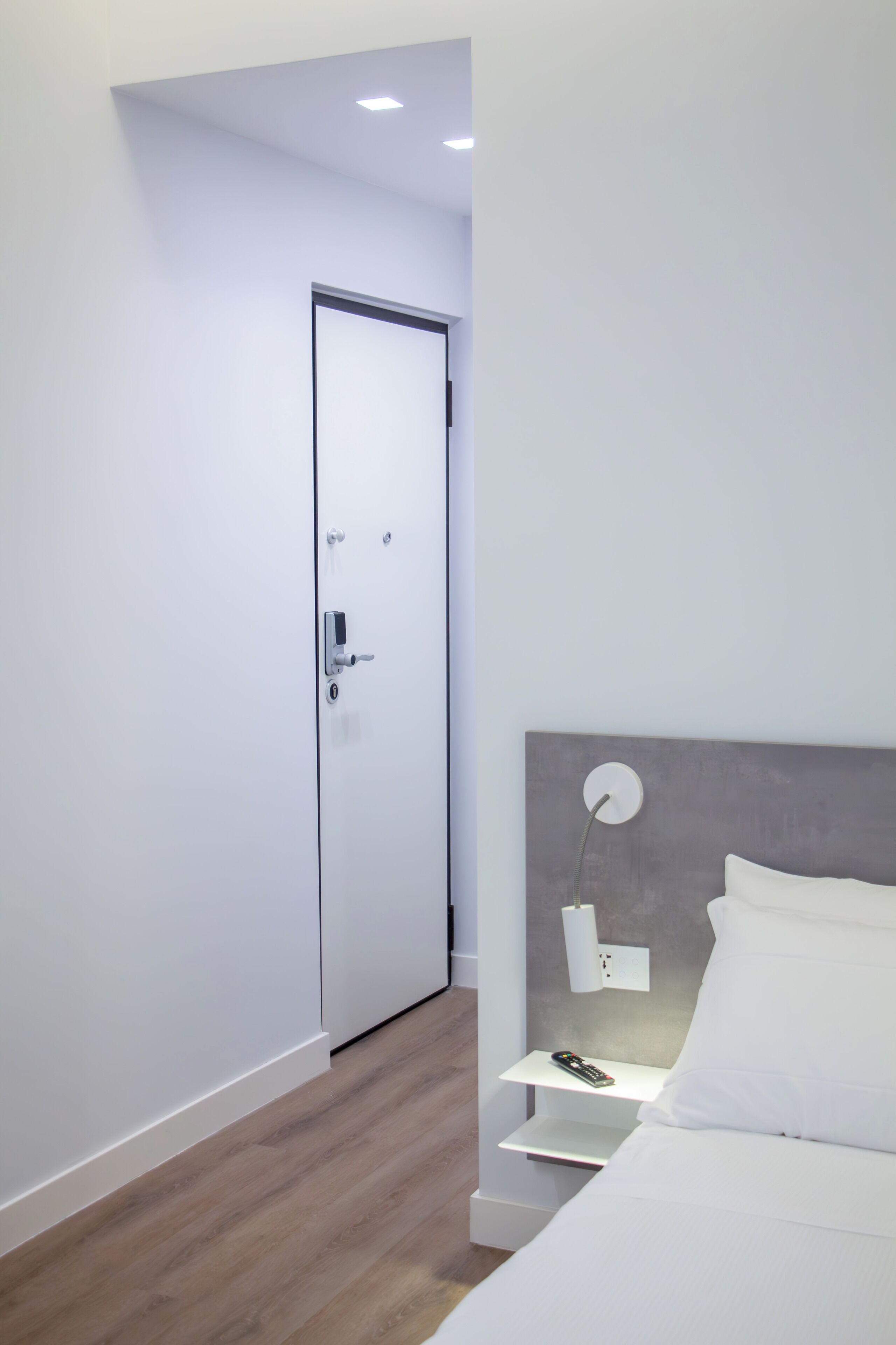A modern bedroom with a white door, a bedside table with a lamp, and a bed with white bedding.