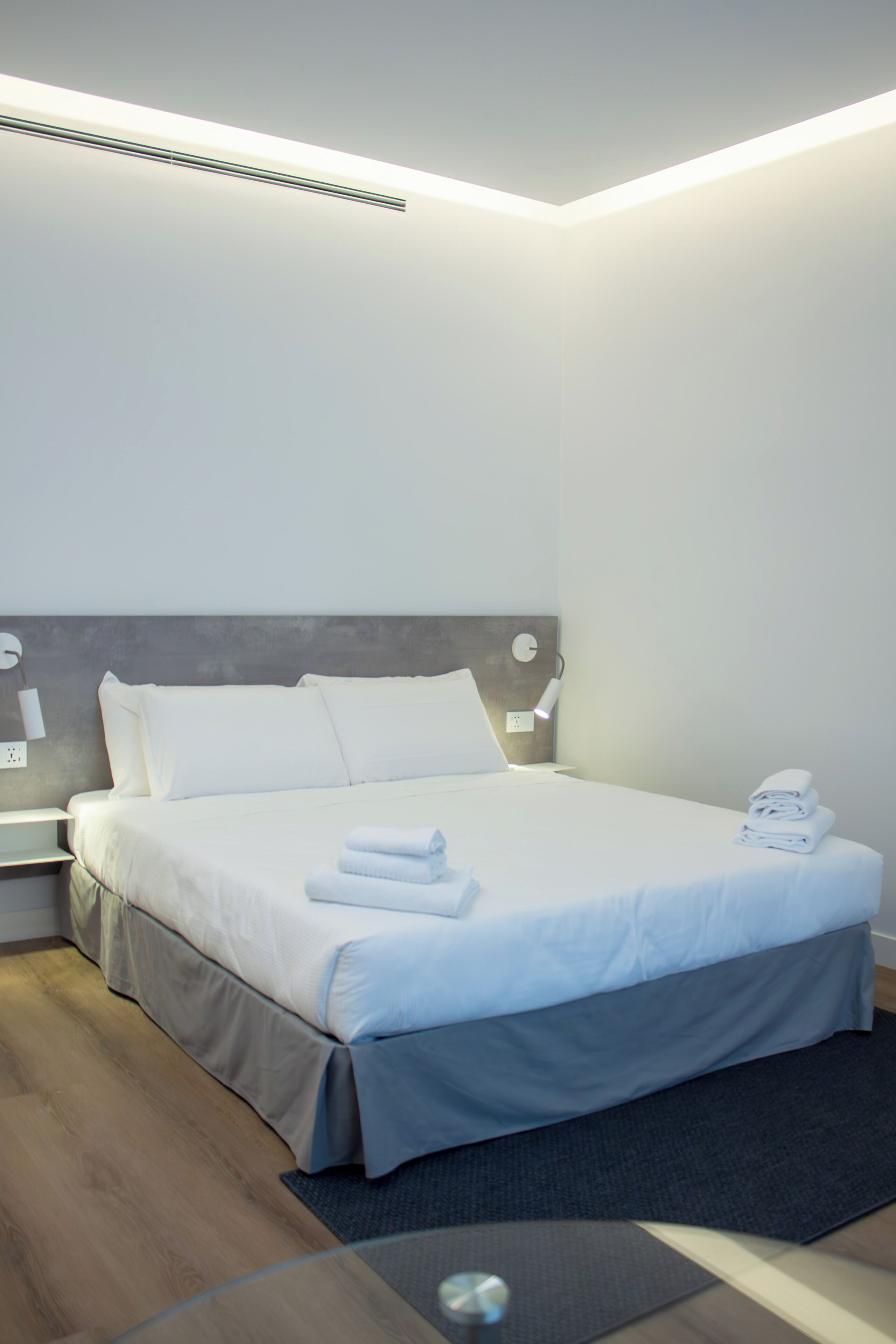 A modern hotel room with a neatly made bed, a bedside table with a lamp, and a power outlet on the wall.