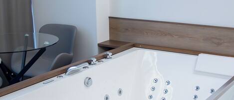 A modern hotel room with a large white bathtub, a glass table, and a wooden headboard.