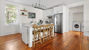 Cocina moderna con armarios blancos, suelo de madera, una isla central con taburetes de bar, electrodomésticos de acero inoxidable y un amplio ventanal con persianas.