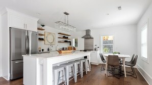 Una cocina moderna con un refrigerador de acero inoxidable, armarios blancos, una isla central con taburetes, una mesa de comedor con sillas y un amplio ventanal con persianas.
