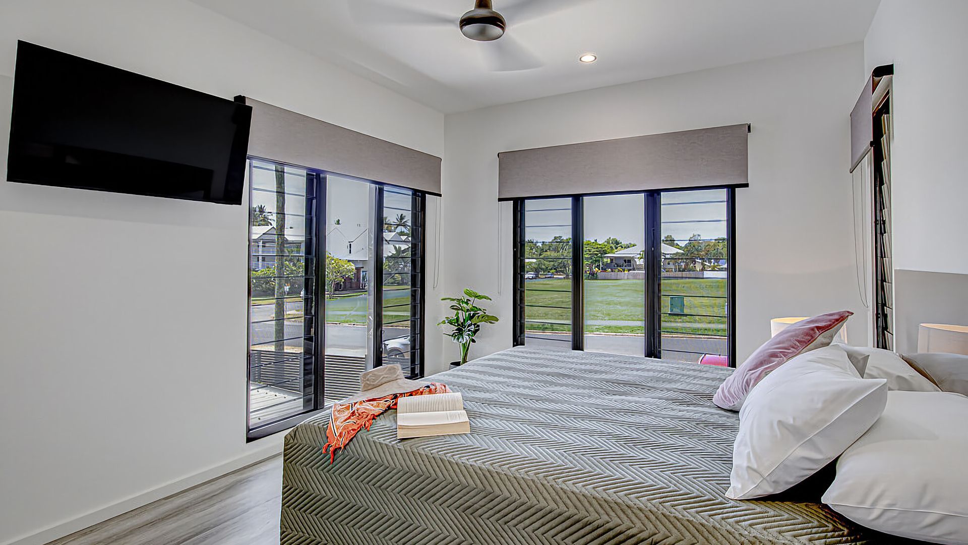 A bedroom with a bed, a TV mounted on the wall, and large windows offering a view of the outdoors.
