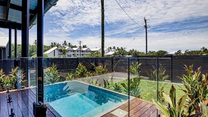 Une piscine dotée d’une terrasse en bois, entourée de plantes tropicales et délimitée par une clôture en métal.