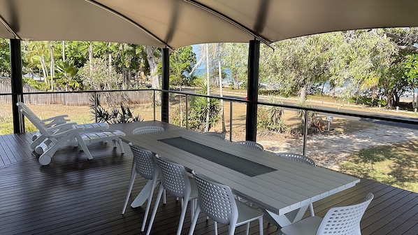 An outdoor patio with a wooden table and white chairs, surrounded by a covered area with glass railings.