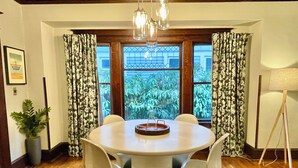 A dining area with a round table, white chairs, a chandelier, and a view through large windows.