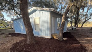 A small, light blue wooden structure with a door and windows, surrounded by mulch and trees. - Shaanti Living • Cozy Studio Blue (Melrose)
