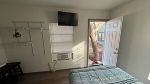 A bedroom with a bed, a TV mounted on the wall, a window with blinds, and a door leading outside. - Shaanti Living • Cozy Studio Blue (Melrose)