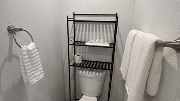 A compact bathroom with a white toilet, a metal shelf unit, a white towel rack, a white basket, and a pink bath mat.