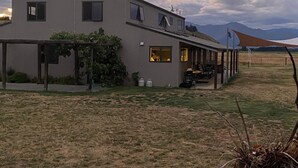 Un edificio moderno de dos plantas con un patio cubierto, un pequeño jardín con hiedra y vistas a las montañas al fondo.
