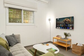 A living room with a sofa, a coffee table, a TV mounted on the wall, and a window with blinds. - Cozy Old North Stay • Steps from Yarkon Park (Tel Aviv)