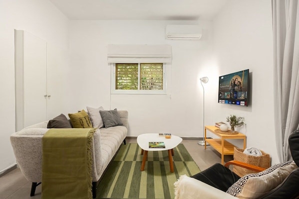 A living room with a sofa, a coffee table, a TV mounted on the wall, and a window with blinds.