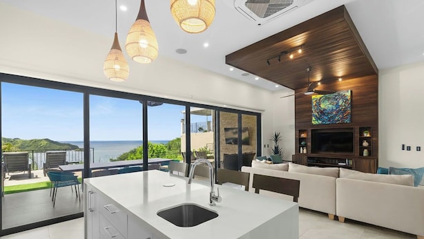 A modern kitchen with a large island, a dining area, and a living room with a TV and artwork.