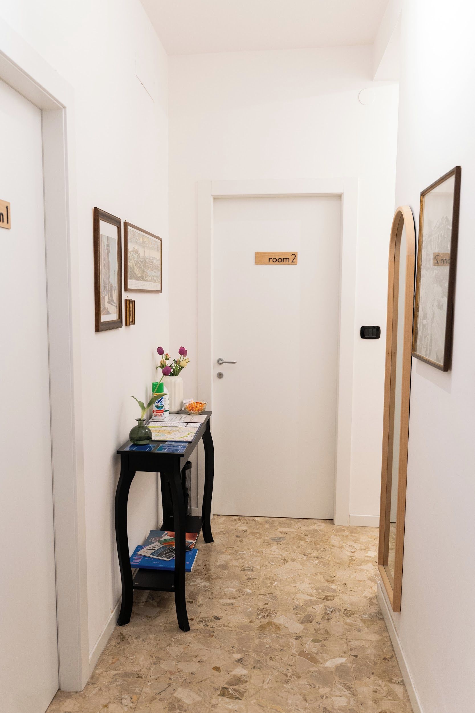Un couloir avec une porte portant l’inscription « salle 2 », ainsi qu’une petite table sur laquelle se trouvent un vase de fleurs et une carte.