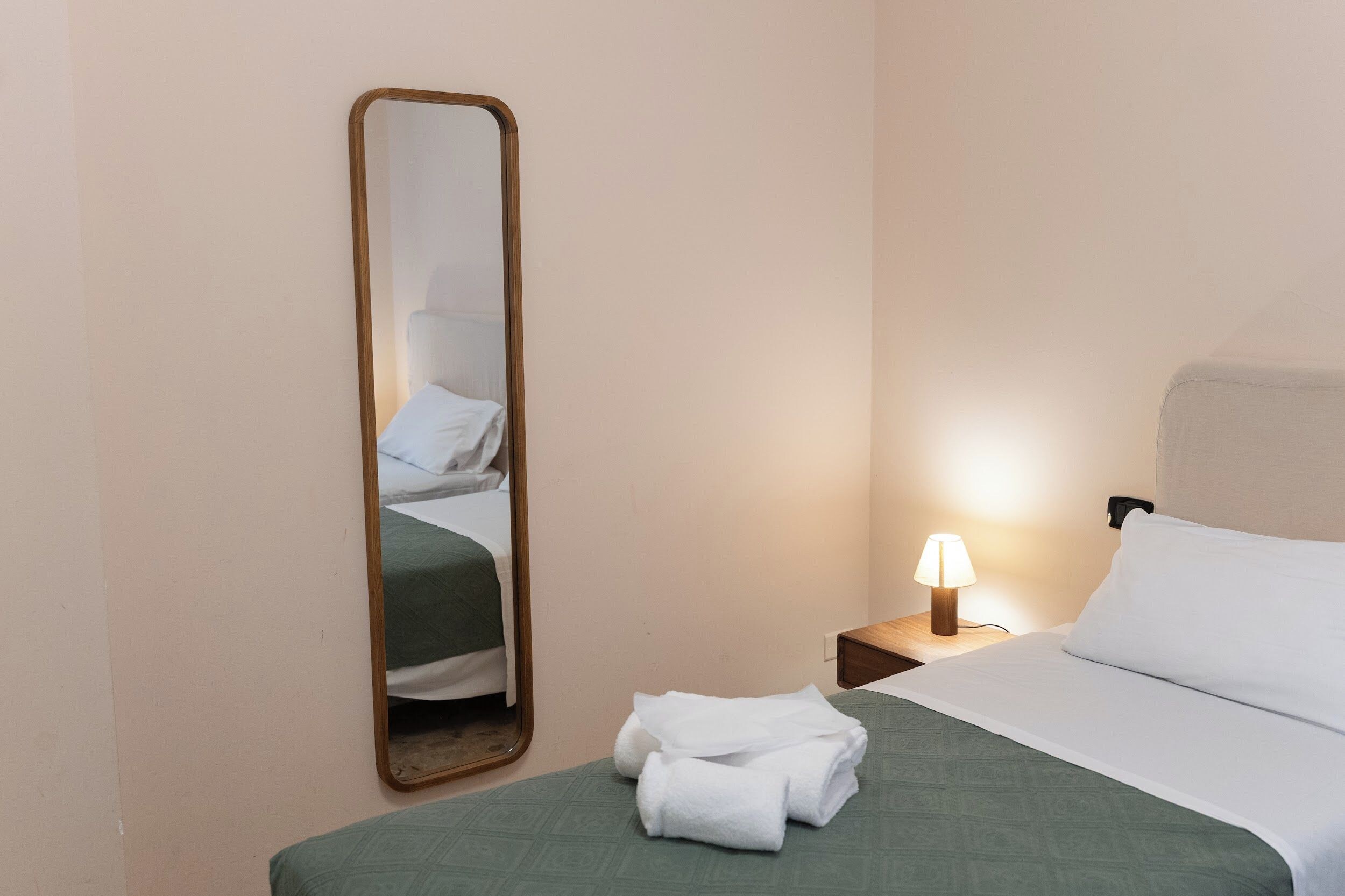 A hotel room with a bed, a nightstand with a lamp, a mirror, and a towel on the bed.