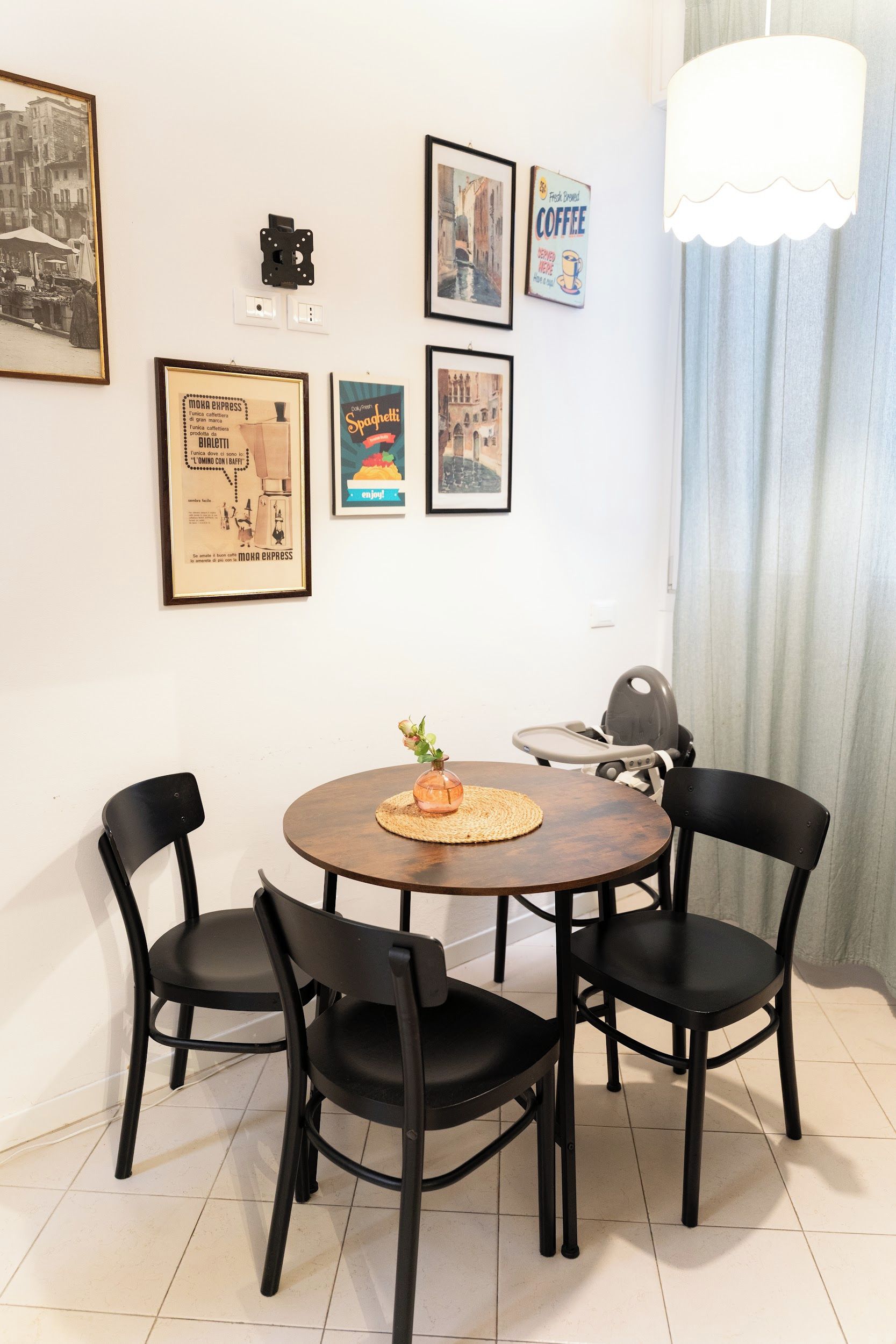 A dining area with a round table, four chairs, and a vase with a flower. There are framed pictures on the wall.