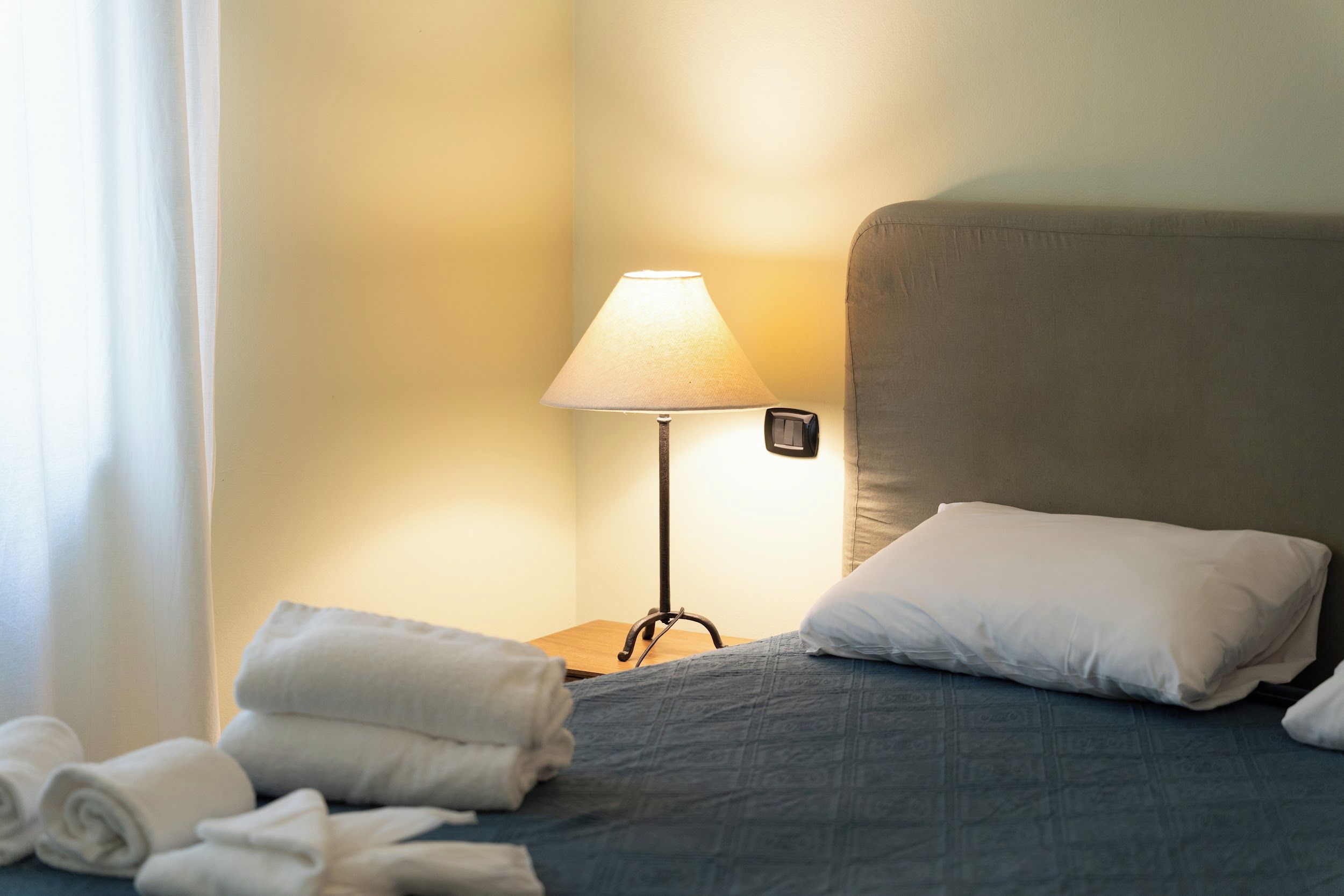 A hotel room with a bed, a lamp, a nightstand, and rolled towels.