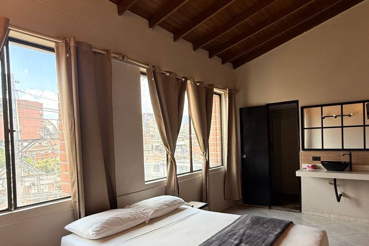 A bedroom with a bed, a nightstand, a window with curtains, a wooden ceiling, and a kitchenette area.