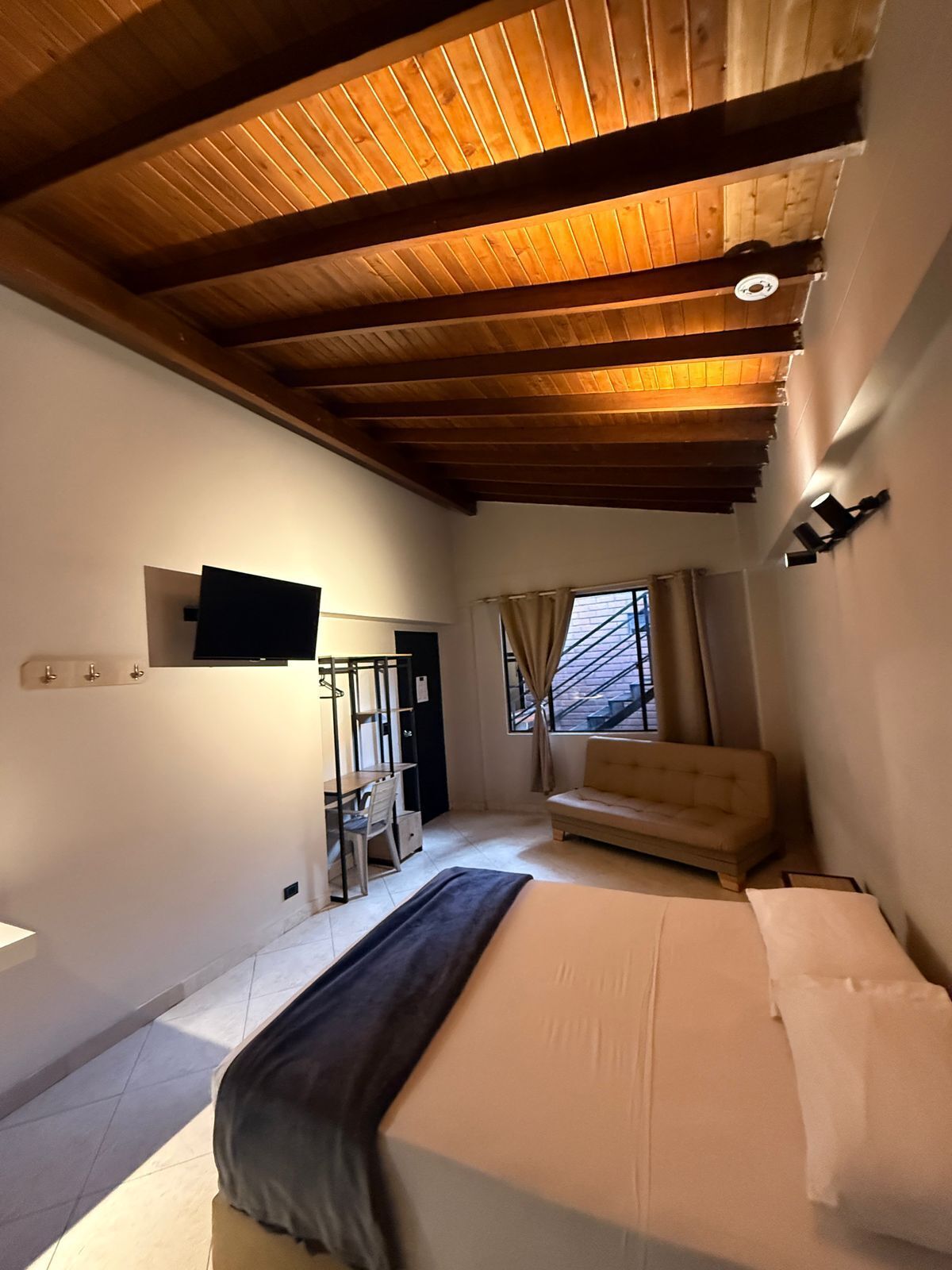 Quarto com cama, escrivaninha, sofá e TV fixada na parede.