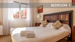 Una cama bien tendida con almohadas y una colcha, una mesita de noche, una lámpara y una ventana con cortinas.