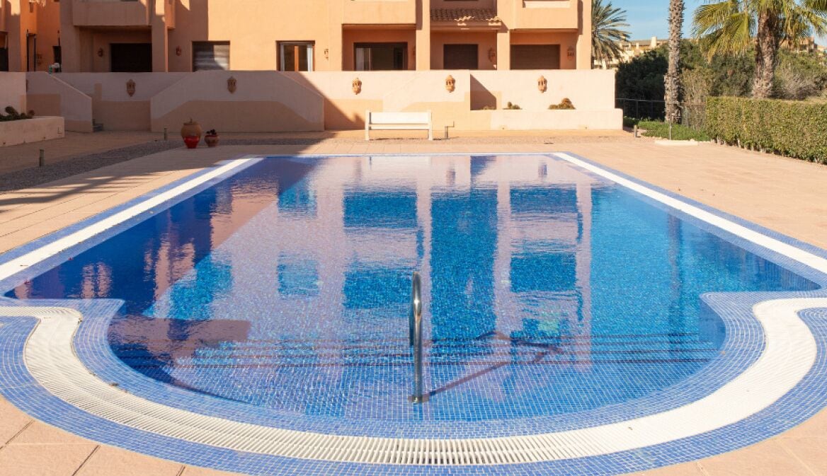 Una piscina con bordo piastrellato, circondata da un edificio beige con balconi, palme e cielo sereno.