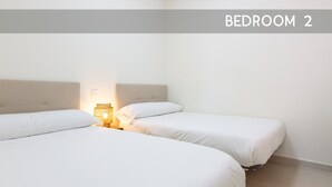 A bedroom with two beds, a nightstand with a lamp, and a plain white wall.