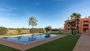 A swimming pool with a surrounding lawn, palm trees, and a building with multiple balconies.