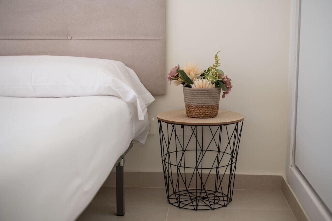 A bed with white linens, a round side table with a woven basket containing flowers, and a geometric wire base.