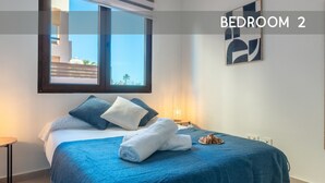 A bedroom with a bed, pillows, a blue blanket, a bedside table with a lamp, and a view through a window.