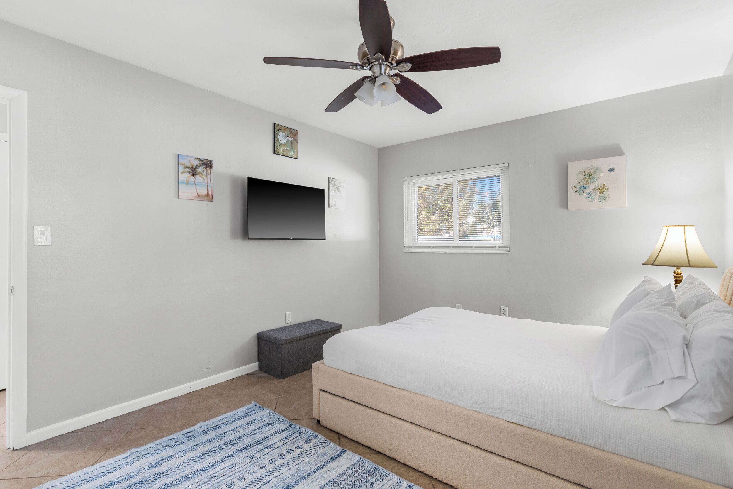 A bedroom with a bed, a ceiling fan, a TV, a window, and a lamp.