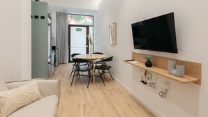 A modern hotel room with a flat-screen TV, a wooden shelf with a vase and a speaker, a dining area with a table and chairs, and a view of the outside through a door.