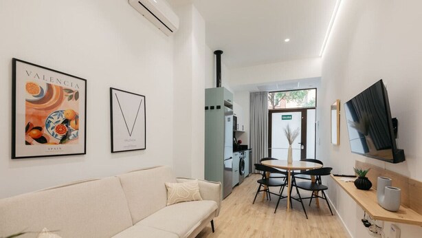 A modern living room with a sofa, a dining table with chairs, a TV mounted on the wall, and a kitchen area visible through an open door.