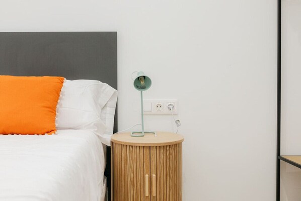 A bed with white and orange pillows, a round wooden nightstand with a green lamp, and a gray headboard. - Elegant Loft in Valencia (València)