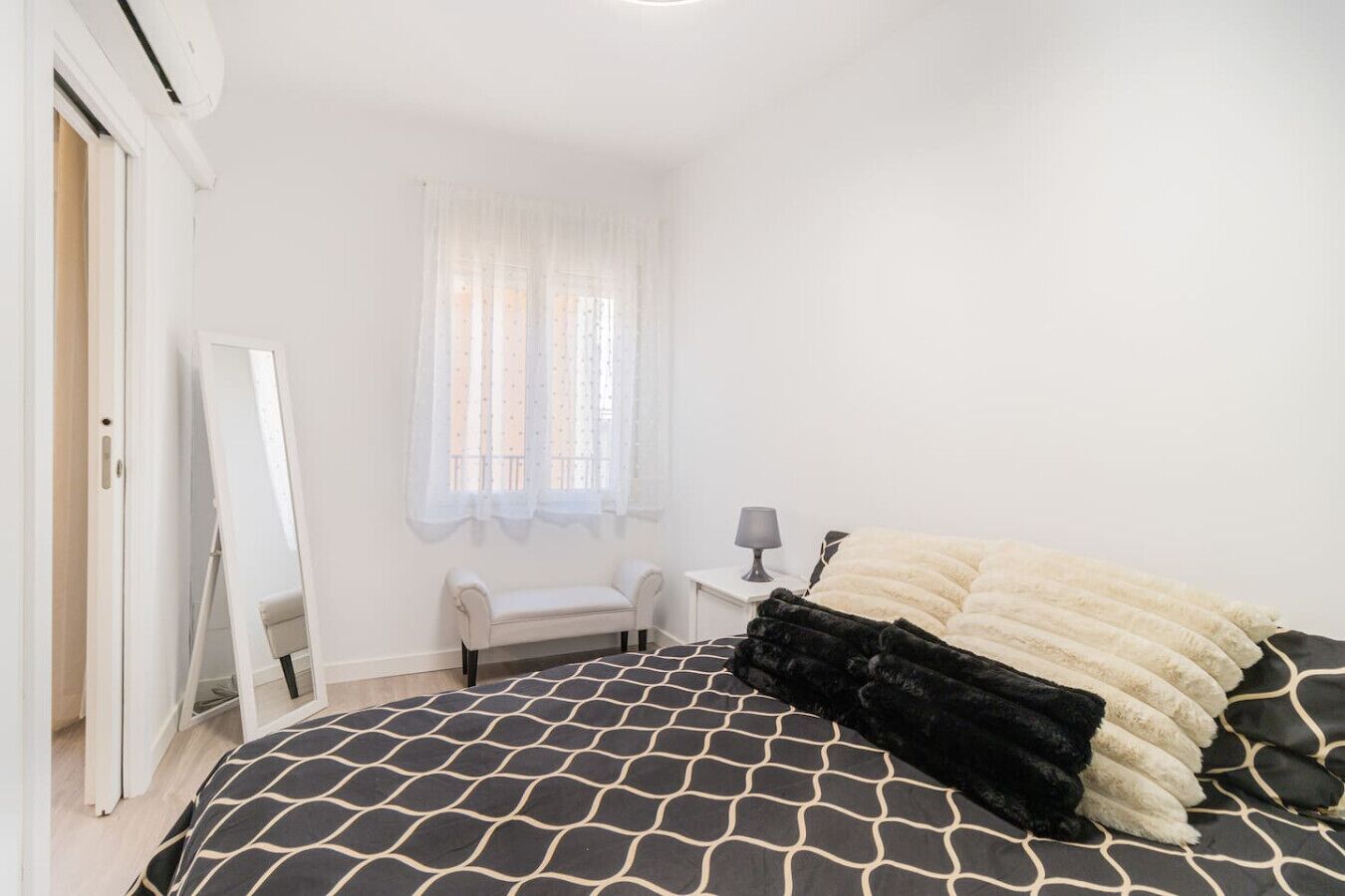 A bedroom with a bed, a chair, a small table with a lamp, and a window with sheer curtains.