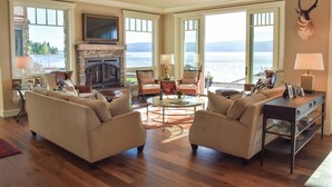 A spacious living room with a fireplace, comfortable seating, and large windows overlooking a lake. - THE FOX HOUSE on Flathead Lake: Lakefront Luxury Living (Bigfork)