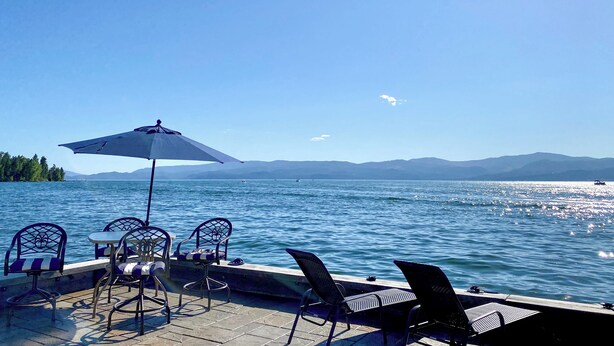 A dock with chairs and an umbrella overlooking a calm lake with boats and mountains in the distance. - THE FOX HOUSE on Flathead Lake: Lakefront Luxury Living (Bigfork)