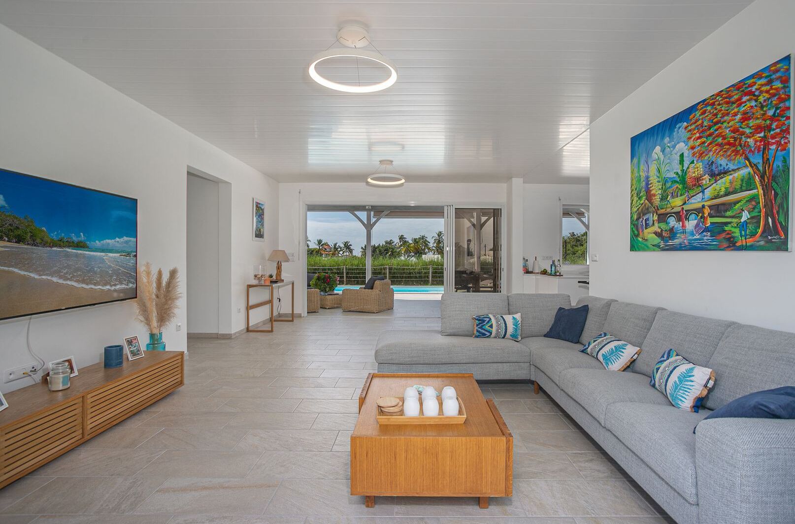 A modern living room with a large flat-screen TV, a grey sofa, a wooden coffee table, and a view of a swimming pool through glass doors.