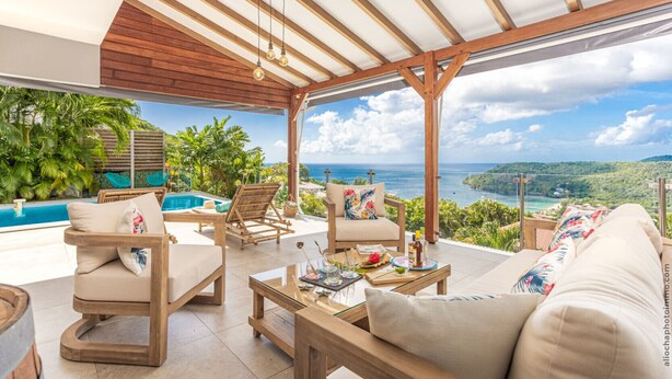 A spacious outdoor lounge area with a pool, wooden furniture, and a scenic ocean view.