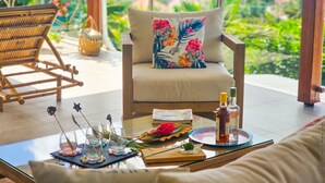 A cozy seating area with a cushioned chair and a sofa, a glass table with drinks and a book, and a view of lush greenery.