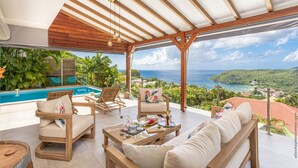 A covered outdoor seating area with a pool, wooden furniture, and a view of the ocean and hills.