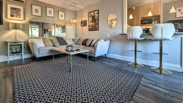 A modern living room with a sofa, coffee table, patterned rug, and a kitchen area visible through an open doorway.