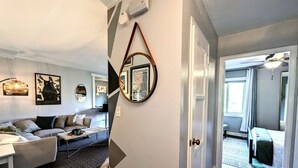 A modern living room with a sofa, coffee table, and decorative mirrors. A door leads to a hallway with a rug and a bench.