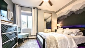 A bedroom with a bed, bedside table, lamp, dresser, and a large window with curtains.