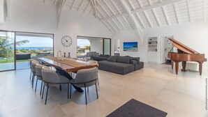A spacious living area with a dining table, sofa, and piano, featuring a high ceiling and large windows overlooking a scenic view. - Luna Moona (Le Vauclin)