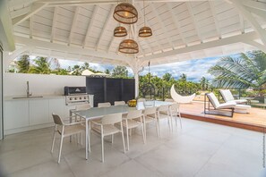 Relax in this outdoor patio with a grill station, dining table, and comfortable seating area overlooking a lush tropical garden.