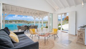 Een moderne woonkamer met een grijze bank, gele kussens en een eethoek met rieten stoelen en een houten tafel. Een grote glazen deur leidt naar een balkon met uitzicht op zee en een wit plafond.