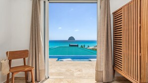 Une pièce avec vue sur une piscine turquoise et l’océan, une chaise en bois et une porte en bois.