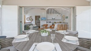 An open-plan living and dining area with a wooden table set for four, a kitchen in the background, and a view of the outdoors. - Blue Cap (Le François)