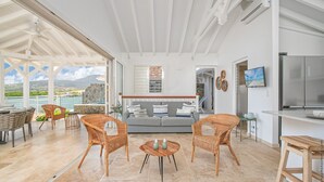 A spacious living area with a view of the sea, a grey sofa, wicker chairs, a wooden coffee table, and a kitchen area with a TV and refrigerator. - Blue Cap (Le François)