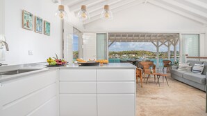 A modern kitchen with a large island, white cabinetry, and pendant lighting. There is a dining area with wicker furniture and a view of the ocean. - Blue Cap (Le François)